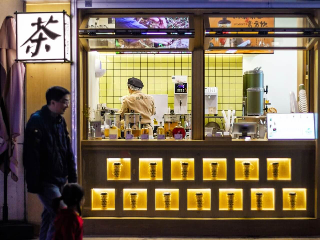 Nighttime bubble tea shop window with illuminated cup displays and a staff member preparing drinks