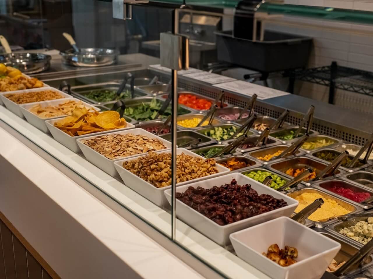 Cafeteria salad bar with bowls of toppings and chilled vegetables behind a glass counter