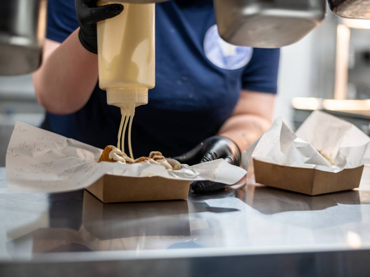 Kitchen worker in gloves drizzling sauce into a paper takeaway tray on a stainless steel counter