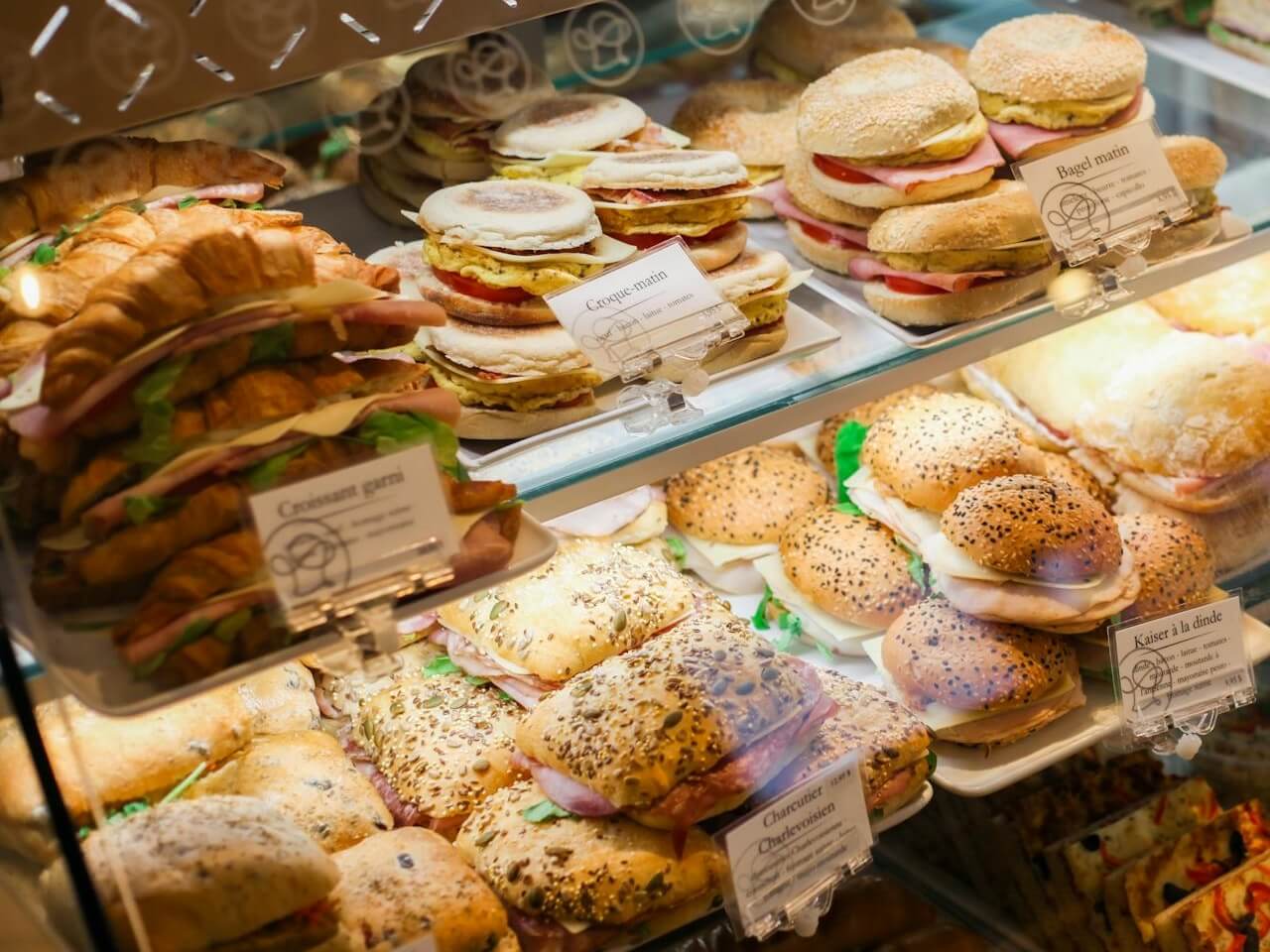 Deli display case filled with croissant sandwiches, bagels, and seeded sandwich rolls on trays
