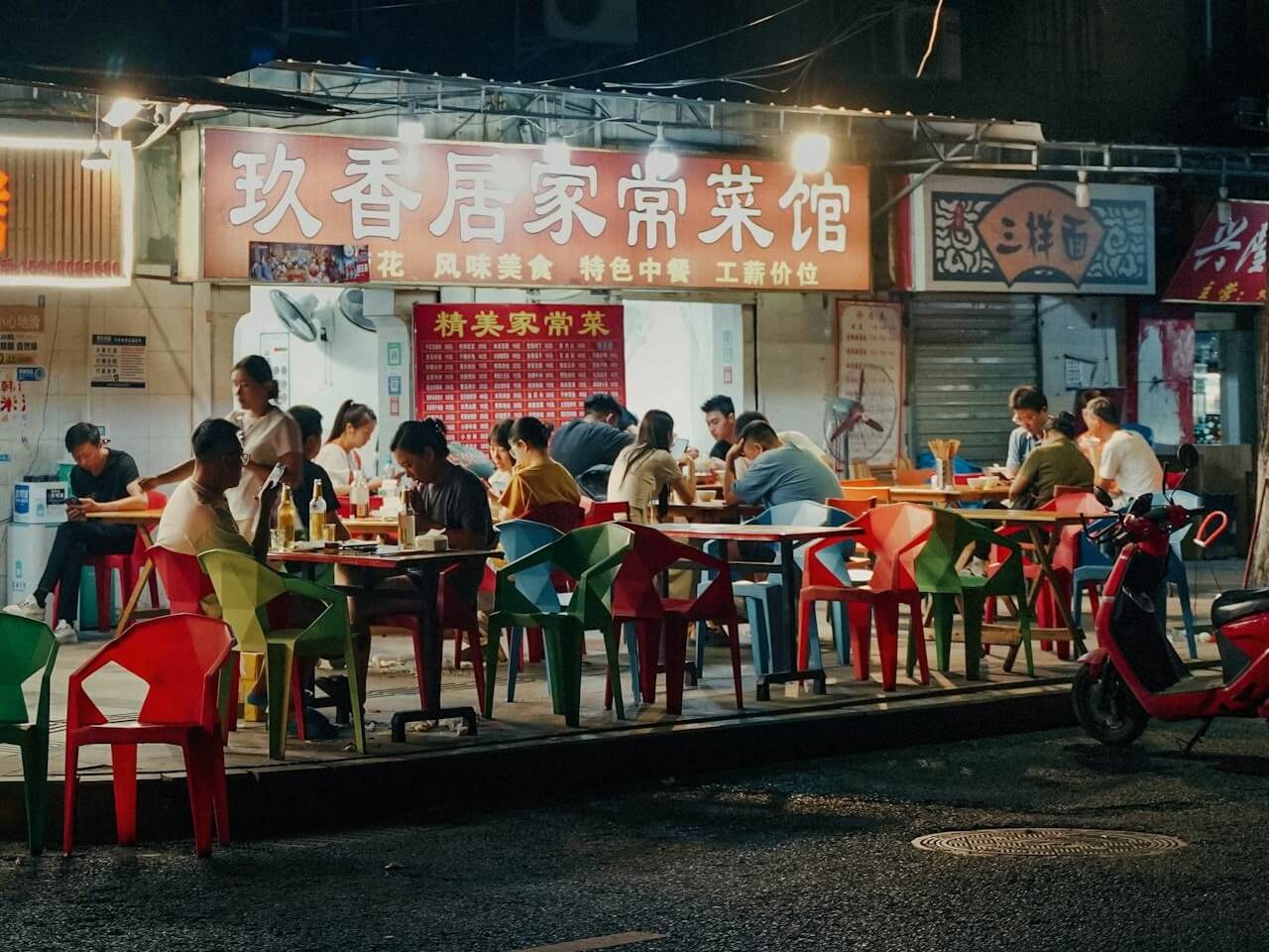 Street-side fast casual restaurant with outdoor seating and diners eating under a bright storefront sign at night