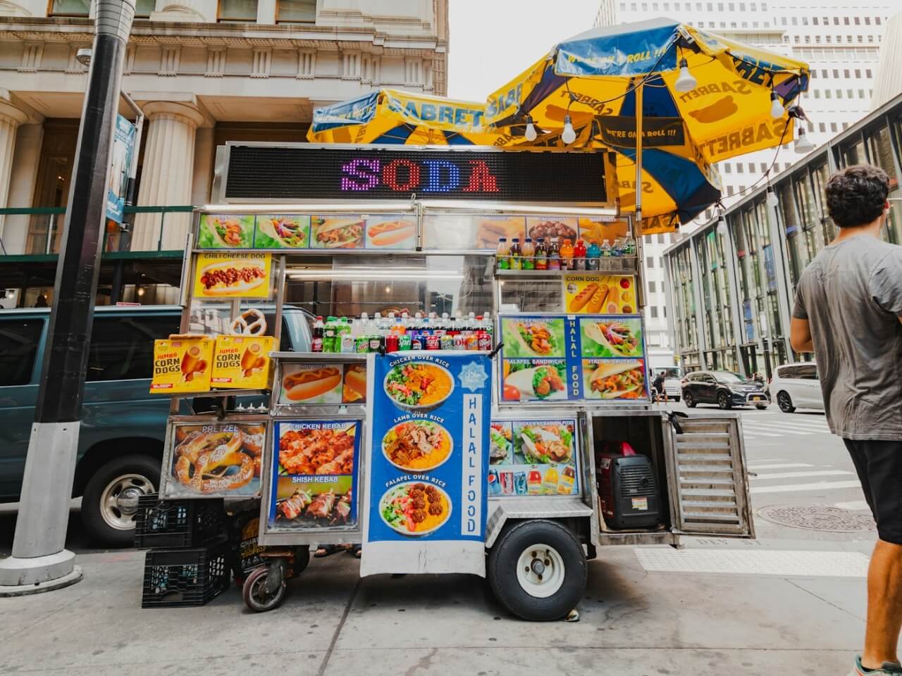 Street food cart with menu photos, a soda sign, and yellow umbrellas on a downtown sidewalk