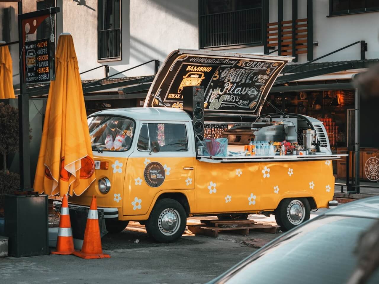 Yellow food truck with an open serving hatch and chalkboard menu parked beside a sidewalk cafe