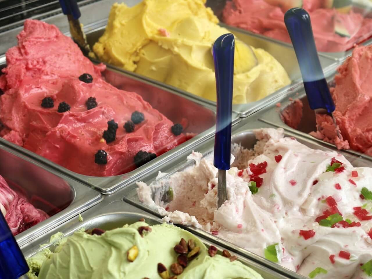 Gelato display case filled with pink, yellow, white, and pistachio flavors in metal tubs