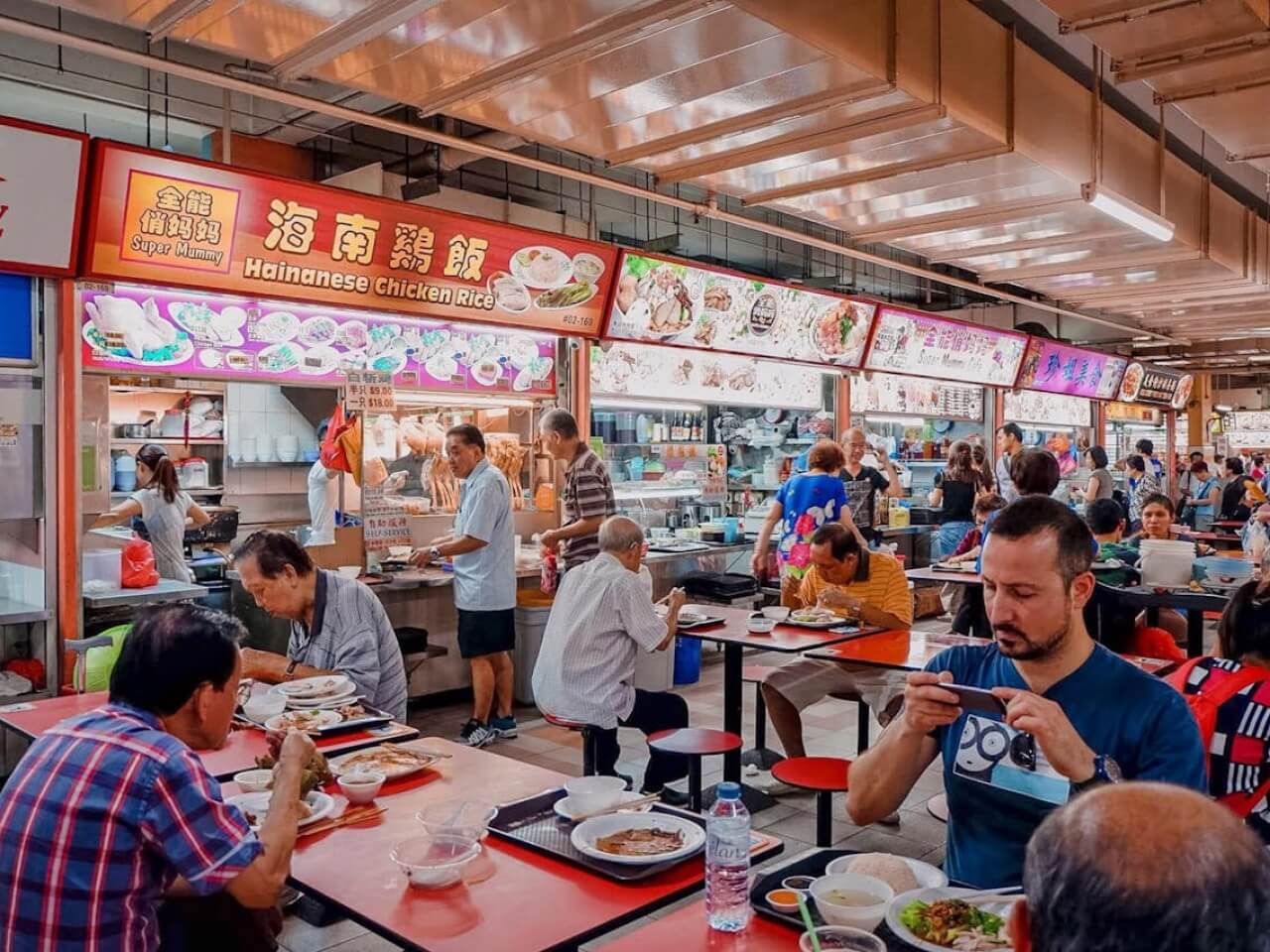 Busy hawker centre with illuminated food stall signs, open counters, and diners seated at red tables