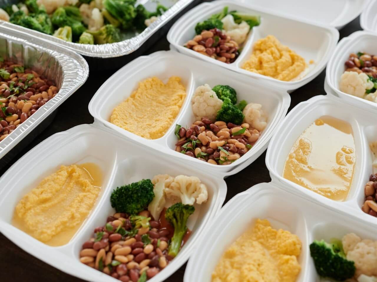 Prepared meal prep containers filled with vegetables, beans, and yellow mash beside catering trays