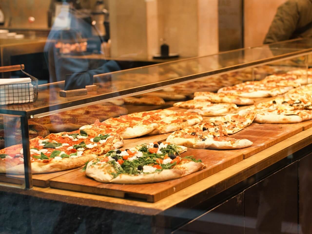 Pizzeria counter display with multiple whole pizzas lined up on wooden boards behind glass