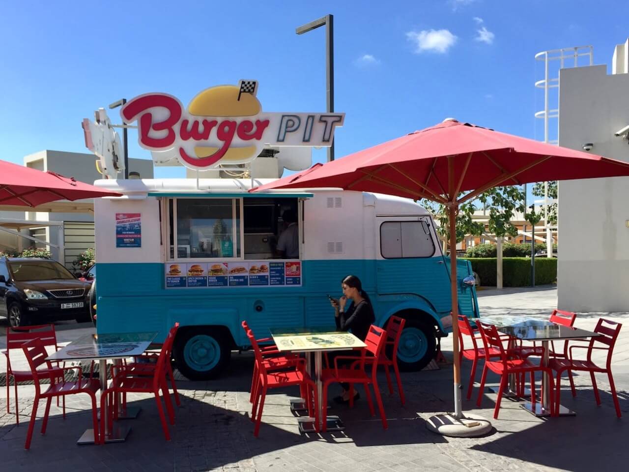Retro burger trailer pop-up with outdoor tables and red umbrellas in a sunny plaza