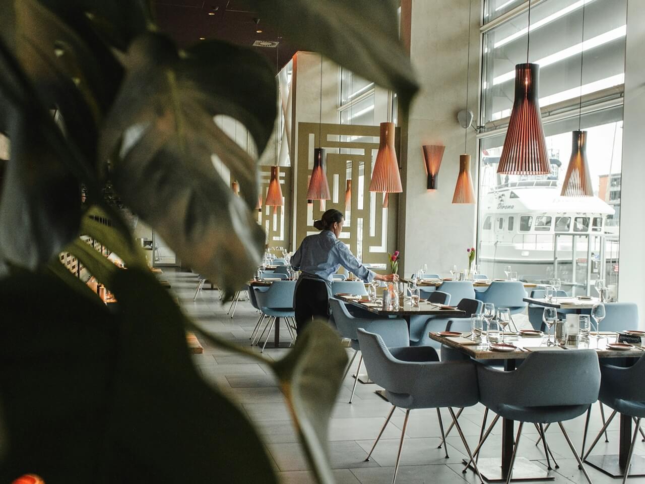 Modern waterfront restaurant dining room with set tables, pendant lights, and a server arranging flowers